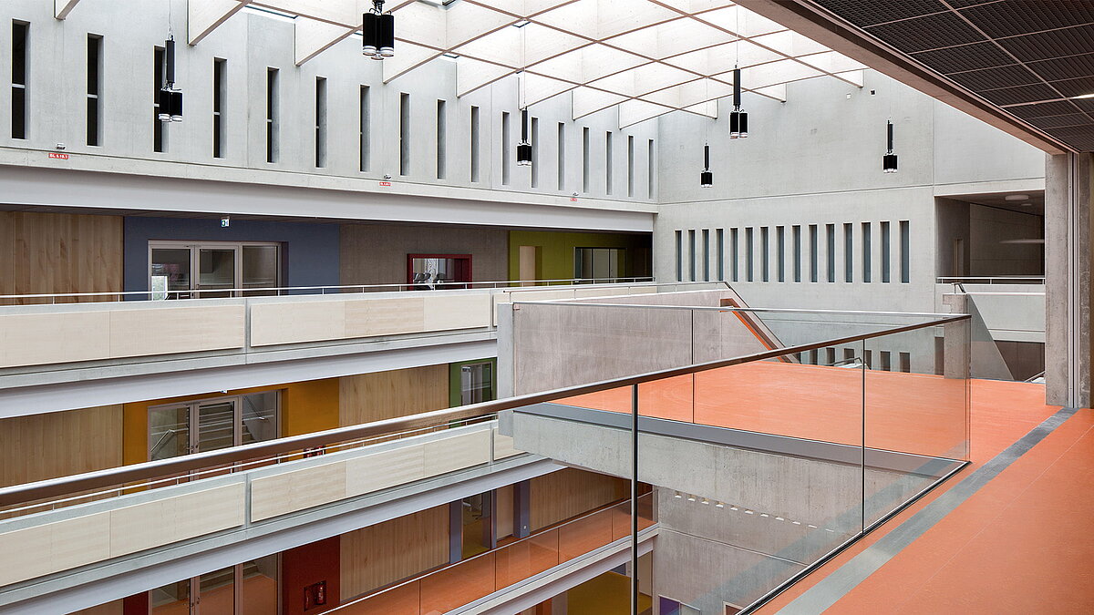 Modern atrium in Willibald-Gluck-Gymnasium in Neumarkt with LAMILUX glass roof, concrete walls, and colorful floors.