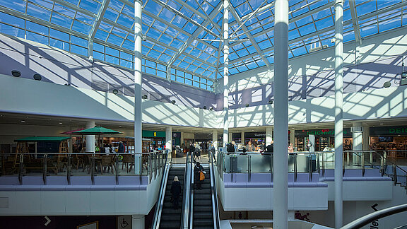 Shopping mall with glass roof, two levels, and people using the escalator