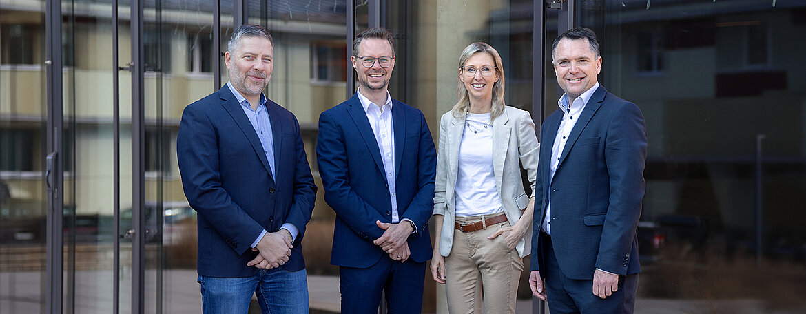 Four people in business attire are standing outside a building, smiling at the camera