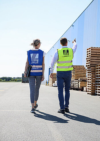 Two people in safety vests walking in front of an industrial building.