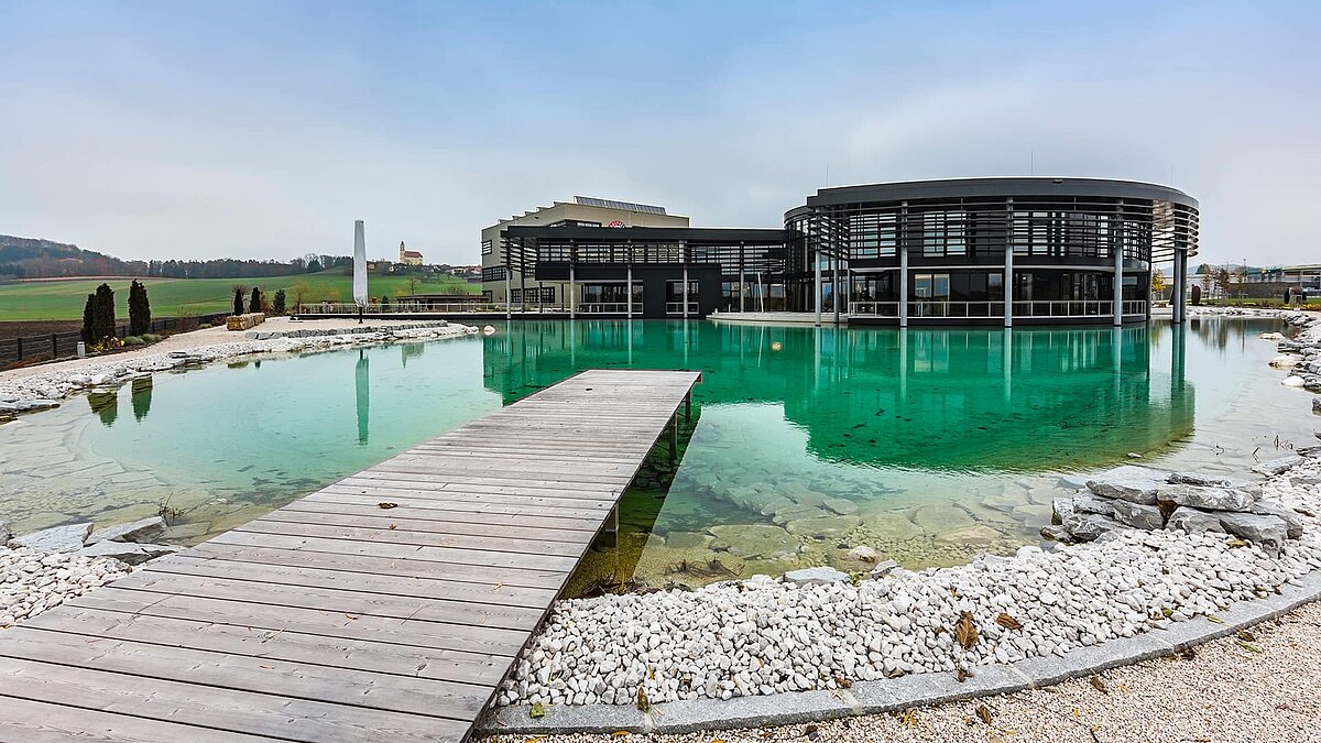 Main building of Zaltech in Moosdorf, Austria, featuring a large pond and a wooden walkway in the foreground.