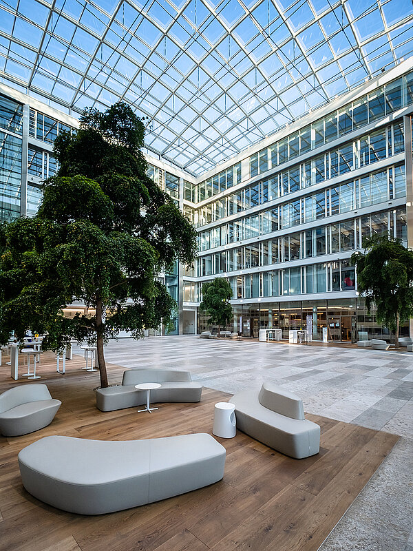 Modern atrium of the Newton office building of the TÜV Süd Group in Munich, featuring a glass roof, large trees, and stylish seating furniture on wooden flooring.