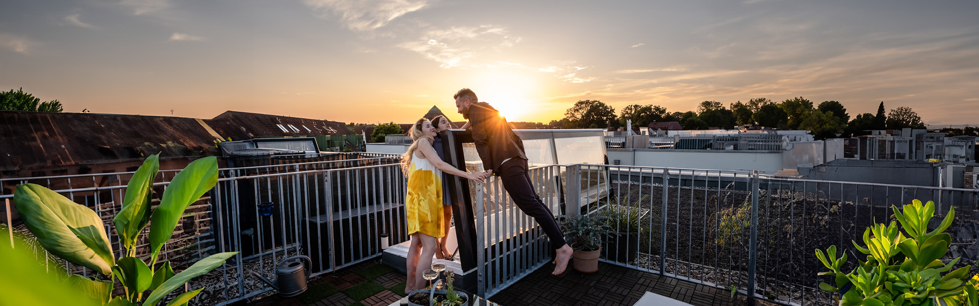 Drei Personen genießen den Sonnenuntergang auf einer Dachterrasse, umgeben von Pflanzen und moderner Architektur