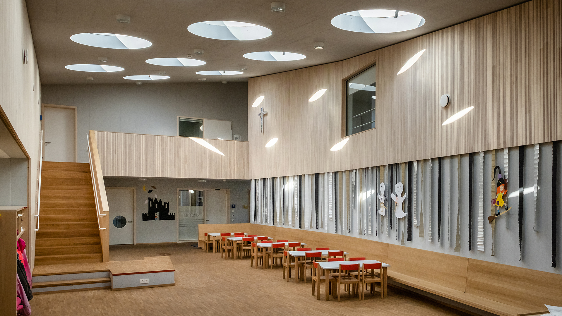 Interior of St. Severin kindergarten with round flat roof windows, wooden walls, and children's tables.