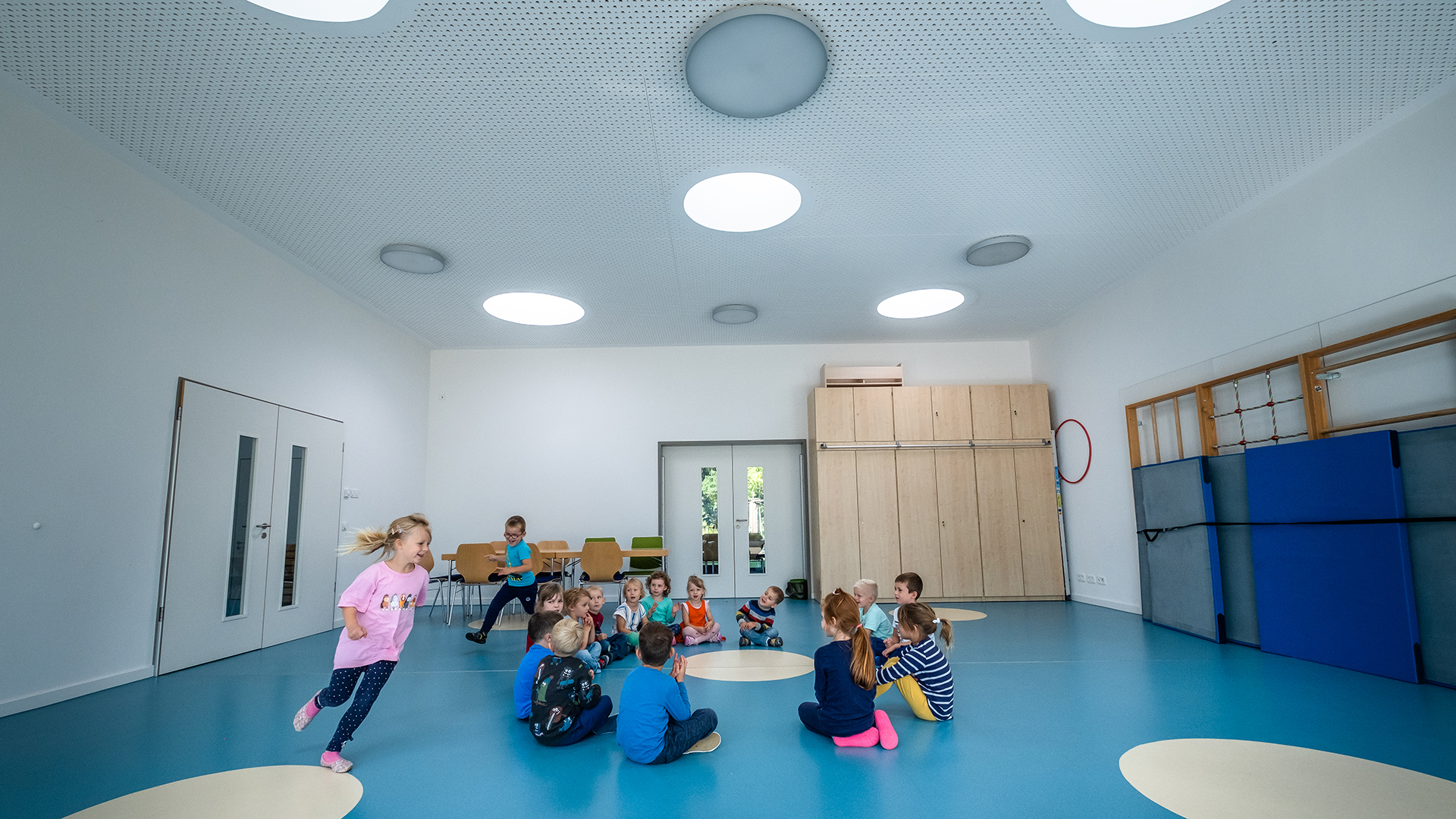 Turnhalle der Kindertagesstätte Sonnenfeld mit spielenden Kindern, blauer Boden und runde Oberlichter.